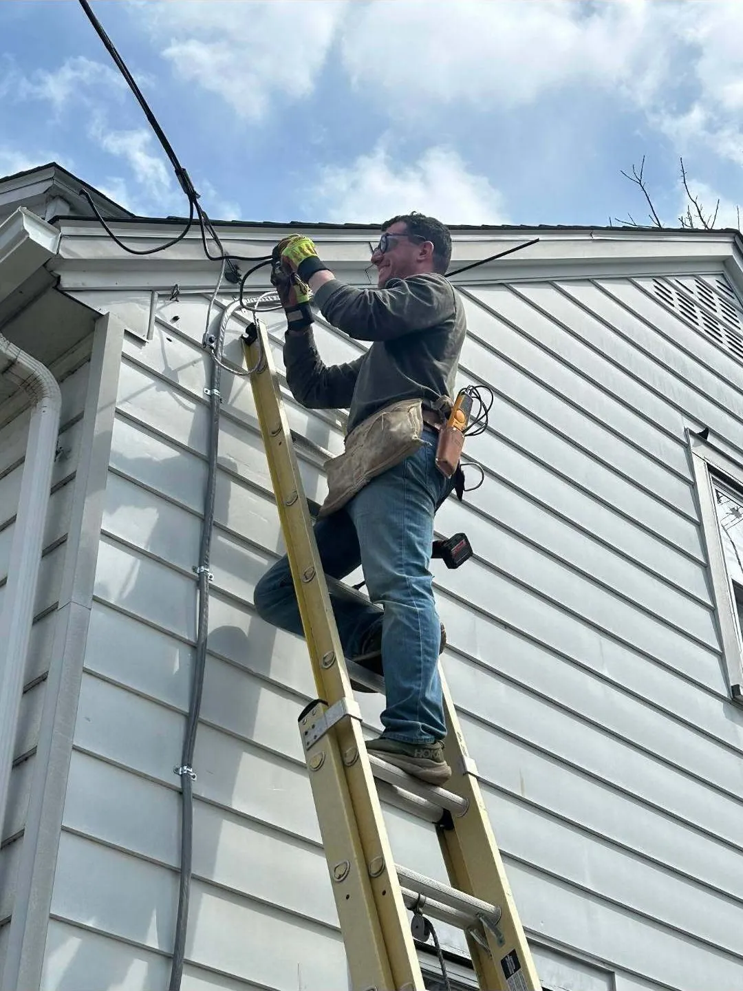 Jim Butler on an extension ladder, connecting overhead service wires at the eave of a house