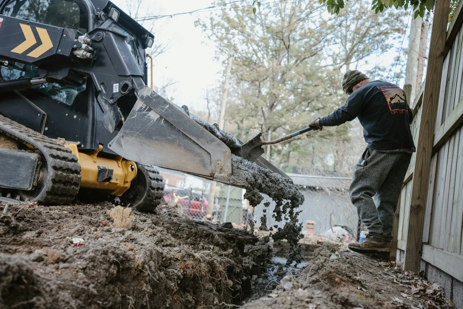 Skid steer digging a trench while a worker shovels alongside it